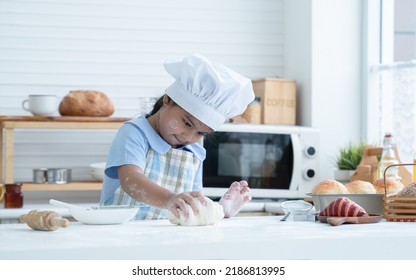 Asian Little Cute Kid Girl Wear Chef Hat And Apron With Flour Mess Up On Face And Arms Have Fun Kneading Dough Bread By Her Hands In Kitchen At Home Or Cooking Class At School. Child Education Concept