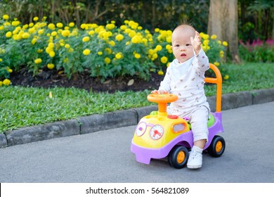 Asian Little Cute Toddler Baby Boy Learn To Ride His First Bike In Park, Kid Drive A Car Toy In Garden With Beautiful Yellow And Pink Flowers, Child Waving Hello Hand Up, Kid First Experience Concept
