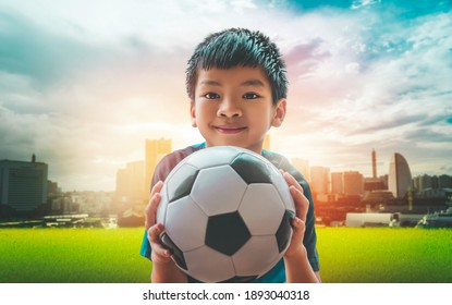 Der kleine asiatische Fußballjunge mit Lächeln hält einen Fußball mit Stadthintergrund.