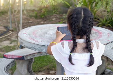 Asian little girl with dark braid is sitting and enjoying playing games on tablet in garden outdoors at home.