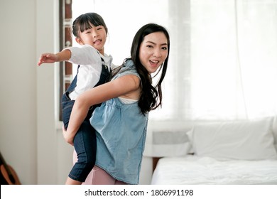 Asian Little Girl Has Fun With Playing On Her Mother Back In Bedroom With Day Light. Concept Of Enjoy With Family Time Of Different Age Of Member In Their House.