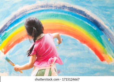 Asian Little Girl Is Painting The Colorful Rainbow And Sky On The Wall And She Look Happy And Funny, Concept Of Art Education And Learn Through Play Activity For Kid Development.