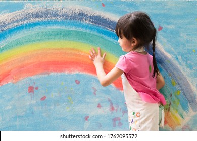 Asian Little Girl Is Painting The Colorful Rainbow And Sky On The Wall And She Look Happy And Funny, Concept Of Art Education And Learn Through Play Activity For Kid Development.