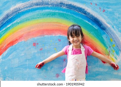 Asian Little Girl Is Painting The Colorful Rainbow And Sky On The Wall And She Look Happy And Funny, Concept Of Art Education And Learn Through Play Activity For Kid Development.