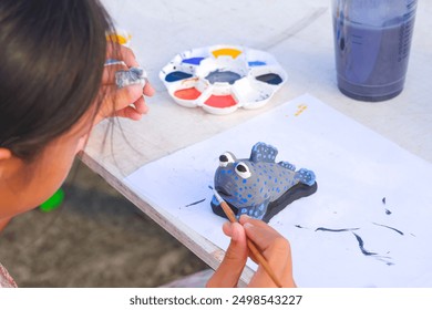 Asian little girl is painting stucco doll in Mudskipper fish shape with watercolors in outdoors walking street market, selective focus - Powered by Shutterstock