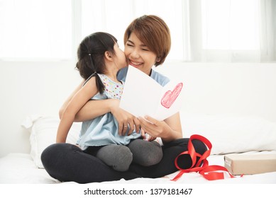 Asian Little Kid Girl Making Surprise With Drawing Red Heart In Postcard Gift For Giving To Her Mother In Bed. Happy Daughter In Blue Cloth Kiss Young Mom Cheek In Mother's Day At Bedroom. 