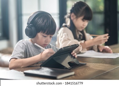 Asian Little Young Children Self Learning At Home. The Boy Using Tablet And Headphones For E-learning At Home. The Girl Doing Homework Writing Note On The Paper Sheet. Education And Technology Concept