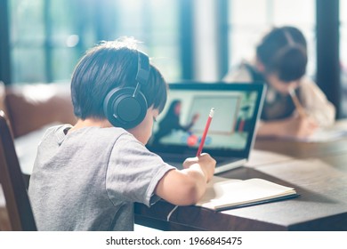 Asian Little Young Children Self Learning At Home. The Boy Using Tablet And Headphones For E-learning At Home. The Girl Doing Homework Writing Note On The Paper Sheet. Education And Technology Concept