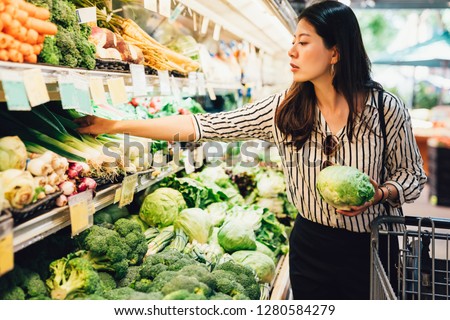 asian local woman buy vegetables and fruits in supermarket. young chinese lady holding green leaf vegetable and picking choosing green onion on cold open refrigerator. elegant female grocery shopping