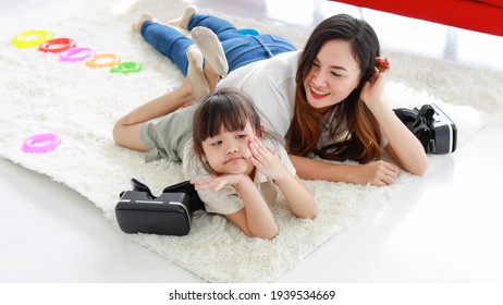 Asian Long Black Hair Mother Wear White Shirt Blue Jeans Socks Smile Lay Down On Fluffy Carpet Together With Bored Face Touchy Daughter After Playing Vr Camera Goggle Game And Colorful Rubber Rings.