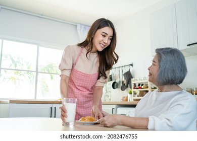 Asian Lovely Family, Young Daughter Prepare Breakfast For Older Mother. Attractive Female Wear Apron Bake Croissant Serve With Milk To Senior Elderly Mom Sitting On Eating Table In Kitchen At House.