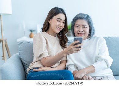 Asian Lovely Family, Young Daughter Use Phone Selfie With Older Mother. Beautiful Girl And Senior Elderly Woman Sit On Sofa, Enjoy Video Call By Internet Online Talking Together In Living Room At Home