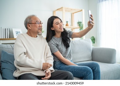 Asian Lovely Family, Young Daughter Use Phone Selfie With Older Father. Beautiful Girl And Senior Elderly Male Sit On Sofa, Enjoy Video Call By Internet Online Talking Together In Living Room At Home.