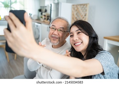 Asian Lovely Family, Young Daughter Use Phone Selfie With Older Father. Beautiful Girl And Senior Elderly Male Sit On Sofa, Enjoy Video Call By Internet Online Talking Together In Living Room At Home.