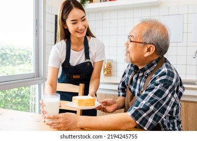 Asian Lovely Family, Young Daughter Prepare Breakfast For Older Father. Attractive Female Wear Apron Bake Bread Serve With Milk To Senior Elderly Dad Sitting On Eating Table In Kitchen At House.