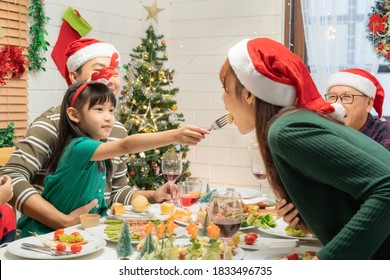 Asian Lovely Girl In Reindeer Headband Feeding Her Mother A Cocktail Sausages Rolls While Having Christmas Dinner Together With Family In A Christmas Decorated Dining Room At Home.