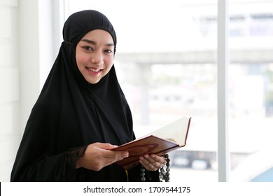 Asian lovely Muslim women nice smailing and holding Quran teaching from god of Islam - Alloh and beads inside mosque room near window. She standing and wearing traditional women clothes - Hijab.