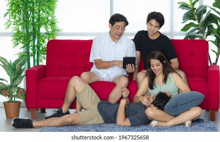 Asian Lovely Warm Family Included Senior Dad Beautiful Mom And Handsome Sons Sitting On Red Sofa And Lay Down On Floor Rest Relax Watching Funny Movie From Tablet Together In Living Room On Weekend.