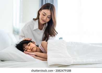 Asian Loving Parents Take Care Of Sleeping Daughter In Bedroom At Home. Attractive Happy Family, Beautiful Mother Puts Blanket On Comfortable Asleep Napping Little Baby Kid On Bed In Morning In House.