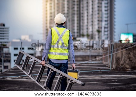 Asian maintenance worker man with safety helmet and green vest carrying aluminium step ladder and tool box at construction site. Civil engineering, Architecture builder and building service concepts