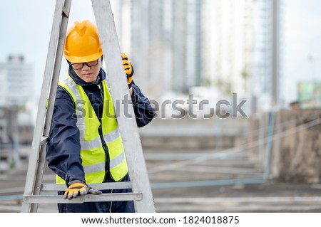 Asian maintenance worker man with safety helmet and reflective suit adjusting aluminium step ladder at construction site. Civil engineering, Architecture builder and building service concepts