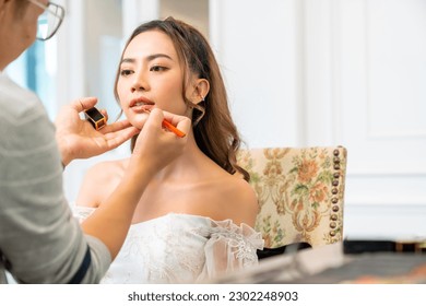 Asian make-up artist applying makeup to beautiful female bride in wedding dress in fitting room at wedding studio. Small business bridal shop owner, beauty salon and marriage ceremony concept. - Powered by Shutterstock