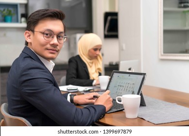 Asian Malay Man Working At Home With Laptop With Colleague At Background