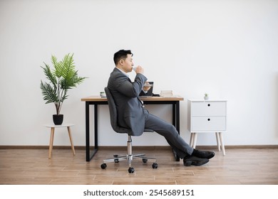 Asian male adult in business attire working on laptop while sipping coffee at desk against white wall. Modern minimalist office setting with potted plant and drawer creating calm atmosphere. - Powered by Shutterstock