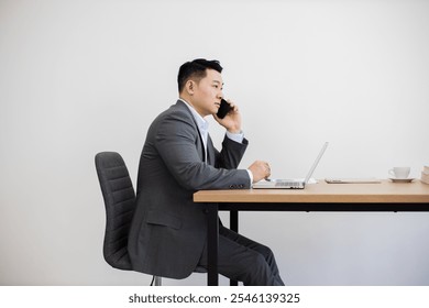 Asian male adult in formal attire working at laptop and making phone call at desk. Modern office setting with laptop, phone, and documents on wooden desk. - Powered by Shutterstock