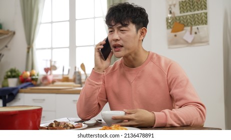 Asian Male Answering Phone While Having Lunch Alone At Home. He Stops Eating And Talks With Unhappy Expression On Face At Dining Table