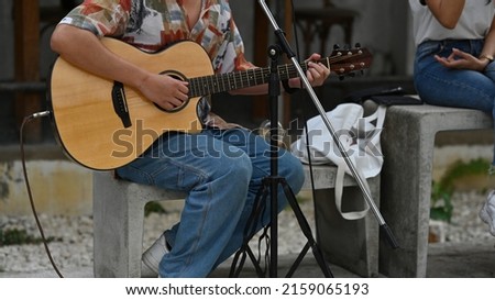 Asian male artist dressed like a hipster playing the guitar next to a microphone standing in the park. Music and abstract concepts.