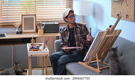Asian male artist painting picture on easel in living room. - Shutterstock ID 2096567755