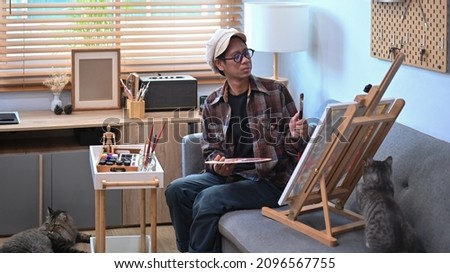 Asian male artist painting picture on easel in living room.