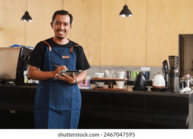 Asian Male business owner at the counter of a coffee shop working using computer tablet, looking at camera - Powered by Shutterstock