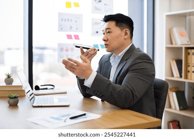 Asian male businessman in modern office sending audio messages via smartphone. He appears focused, demonstrating an engaging work environment. Office features laptop, documents, organized setting. - Powered by Shutterstock