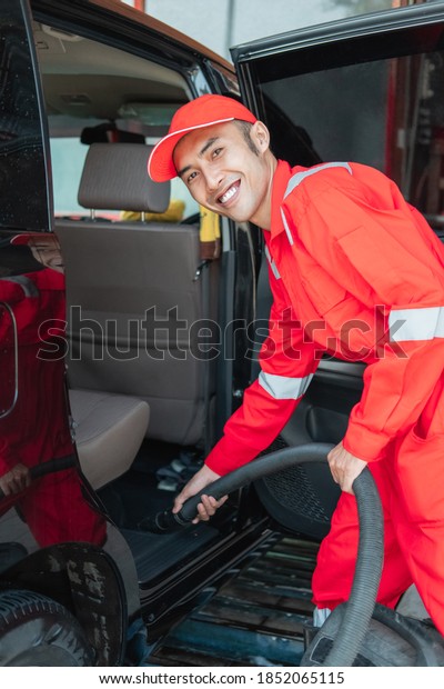 Asian male car cleaner\
wears red smiling uniform while cleaning car floor with vacuum\
cleaner in car salon