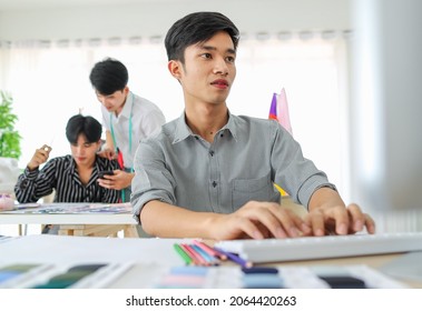 Asian male craftsman browsing data on computer while sitting at table near dressmakers in workshop - Powered by Shutterstock