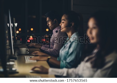 Asian male customer care service with business man smiling and working hard late in night shift at office,call center department,worker and overtime,7 days 24 hour,teamwork with colleagues for success