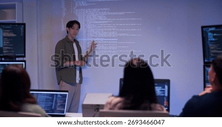 Asian male developer presenting source code to colleagues in a dark tech office. Team listening to coding explanation during software training, AI session, or cybersecurity presentation.