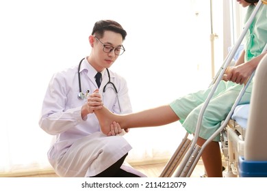 Asian Male Doctor Examining The Ankle Of A Female Patient Sitting On A Nursing Bed She Holds A Cane To Help Her Walk. The Concept Of Ankle Pain, Inflammation, Injury. Physical Therapy