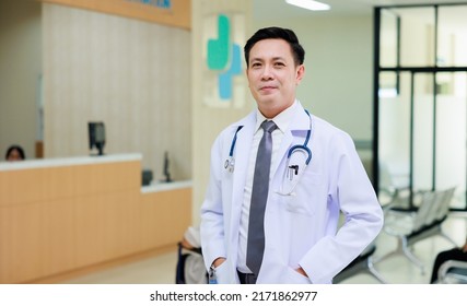 Asian Male Doctor In White Uniform Look At Camera Standing In Patient Reception At Modern Hospital. Healthcare Medical And Medicine Concept.