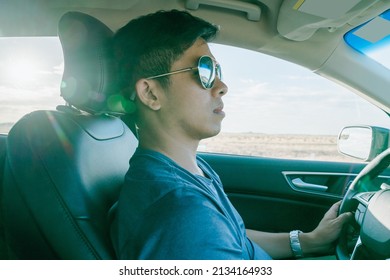 Asian Male Driver Going On A Road Trip On Sunny Day. Japanese Man Wearing Sunglasses Driving Behind Wheel With Dry Roadside View.