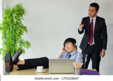 Asian male employees secretly relax, listen to music during working time. He sat put his feet on the desk and is lazy while the employer watched from behind.