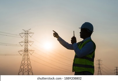 An Asian male engineer using a radio station is working in front of a high voltage pole. In the sunset the sky is beautiful