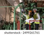 Asian male and female engineers at nuclear power plant standing planning a project with blueprints while large machinery is working behind them, wearing hard hats and vests.