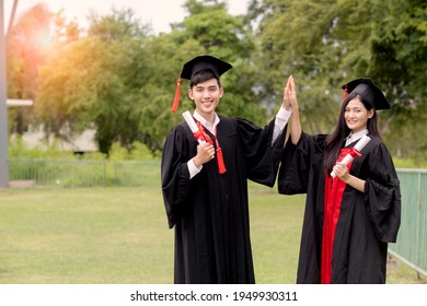 A Asian Male And Female Graduated Student Standing  At Sport Field's College And Hi Five With Smiling And Pround Looking To Camera. Hands Holding Diploma With Triumph.