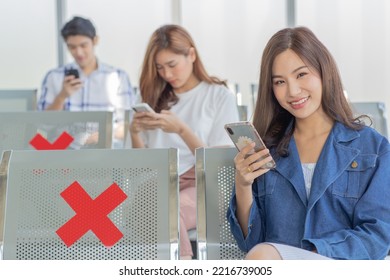 Asian Male And Female Traveler Passengers Sitting Browsing Surfing Internet Via Smartphone On New Normal Social Distancing Chairs With Red Cross Sticker In Airport During Covid19 Pandemic Quarantine.