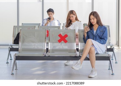 Asian Male And Female Traveler Passengers Sitting Browsing Surfing Internet Via Smartphone On New Normal Social Distancing Chairs With Red Cross Sticker In Airport During Covid19 Pandemic Quarantine.