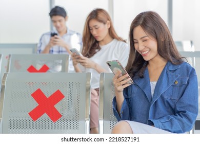 Asian Male And Female Traveler Passengers Sitting Browsing Surfing Internet Via Smartphone On New Normal Social Distancing Chairs With Red Cross Sticker In Airport During Covid19 Pandemic Quarantine.