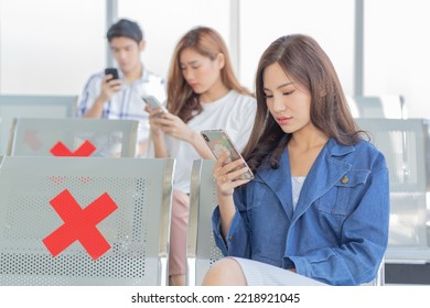 Asian Male And Female Traveler Passengers Sitting Browsing Surfing Internet Via Smartphone On New Normal Social Distancing Chairs With Red Cross Sticker In Airport During Covid19 Pandemic Quarantine.